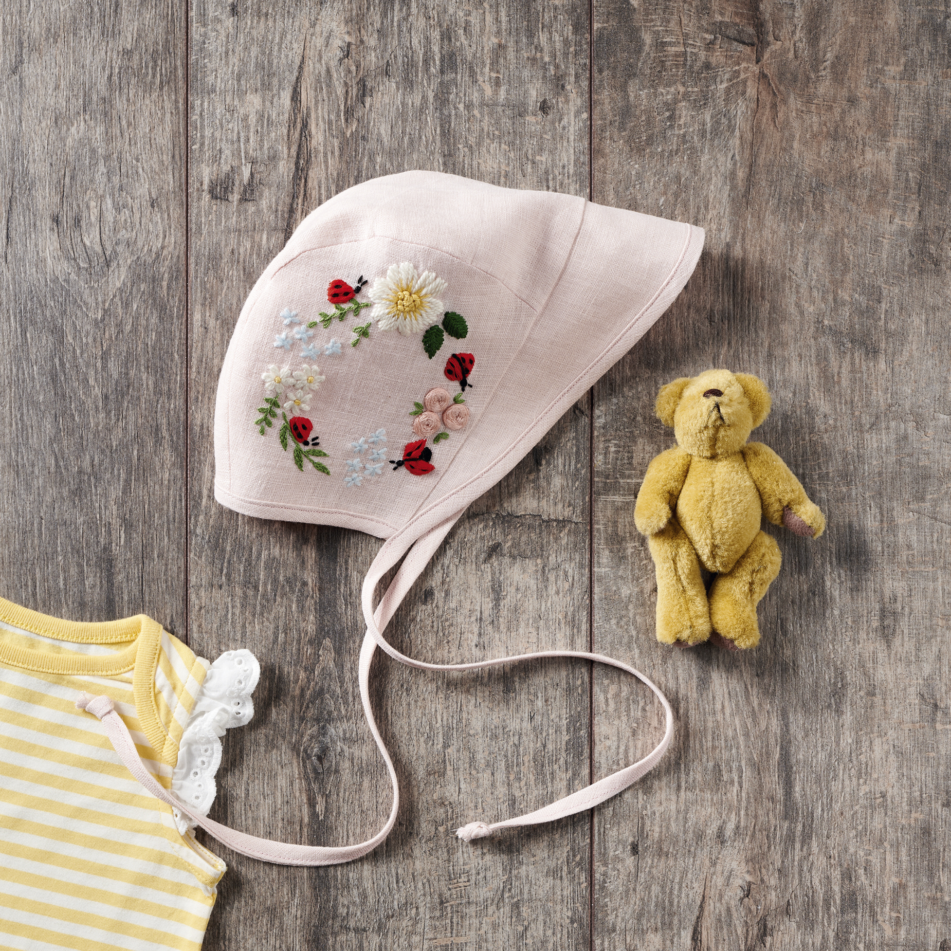 Light pink baby bonnet with ladybird and floral embroidery details. It sits on a dark wood background with a yellow teddy bear next to it and a white and yellow stripy top to the bottom left.