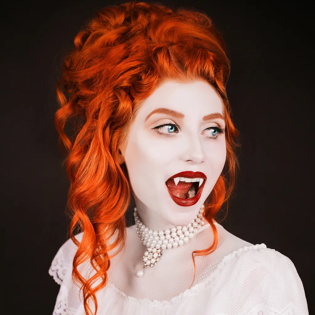 Young vampire woman with long curly hair, pale skin in a white dress on a black background. A beautiful redhead model with red lips. Outfit for halloween. Vampire with open mouth and fangs easy Halloween face paint ideas
