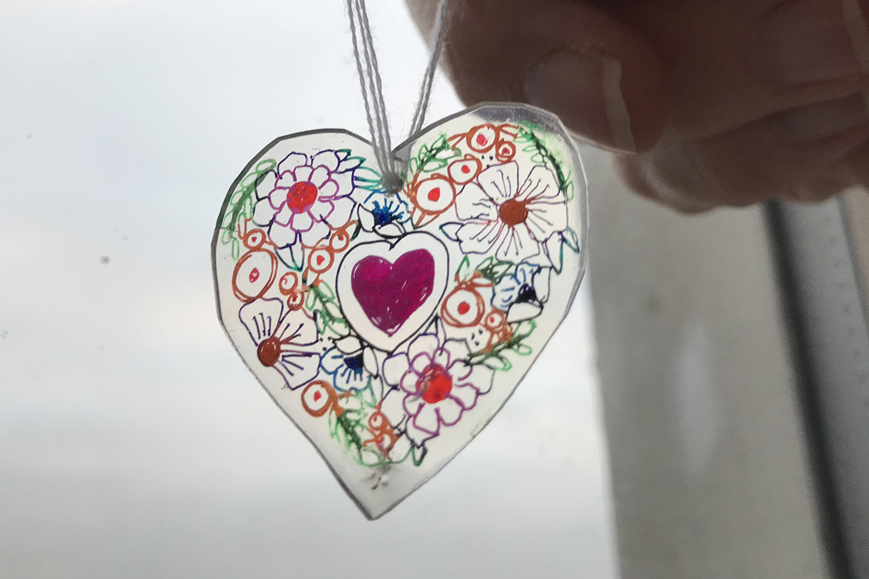 A heart shrinky dink with delicate decorations of hearts, flowers and leaves is held up against the light to show off the detail