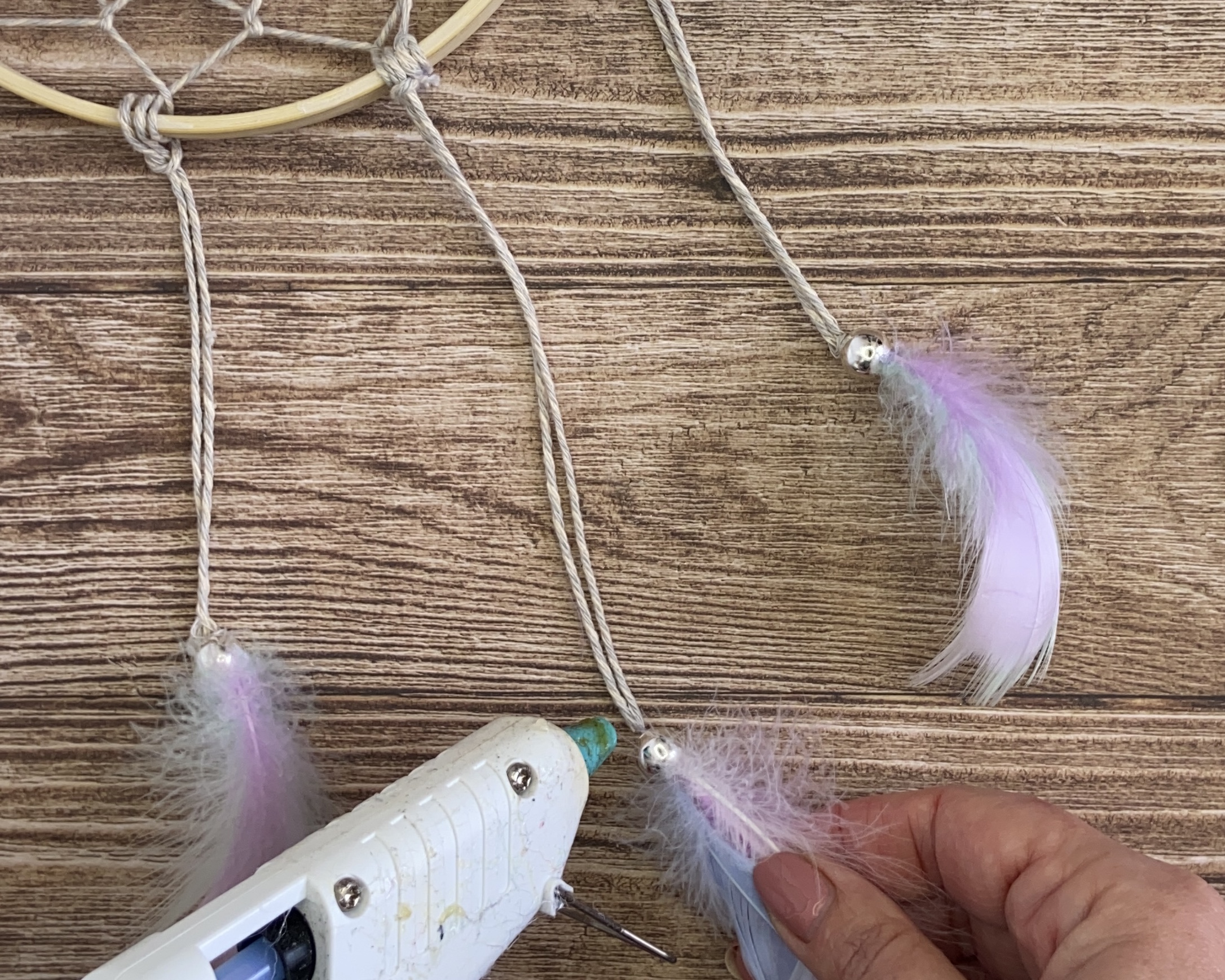 hands using glue gun to attach feather to length of cord under dreamcatcher