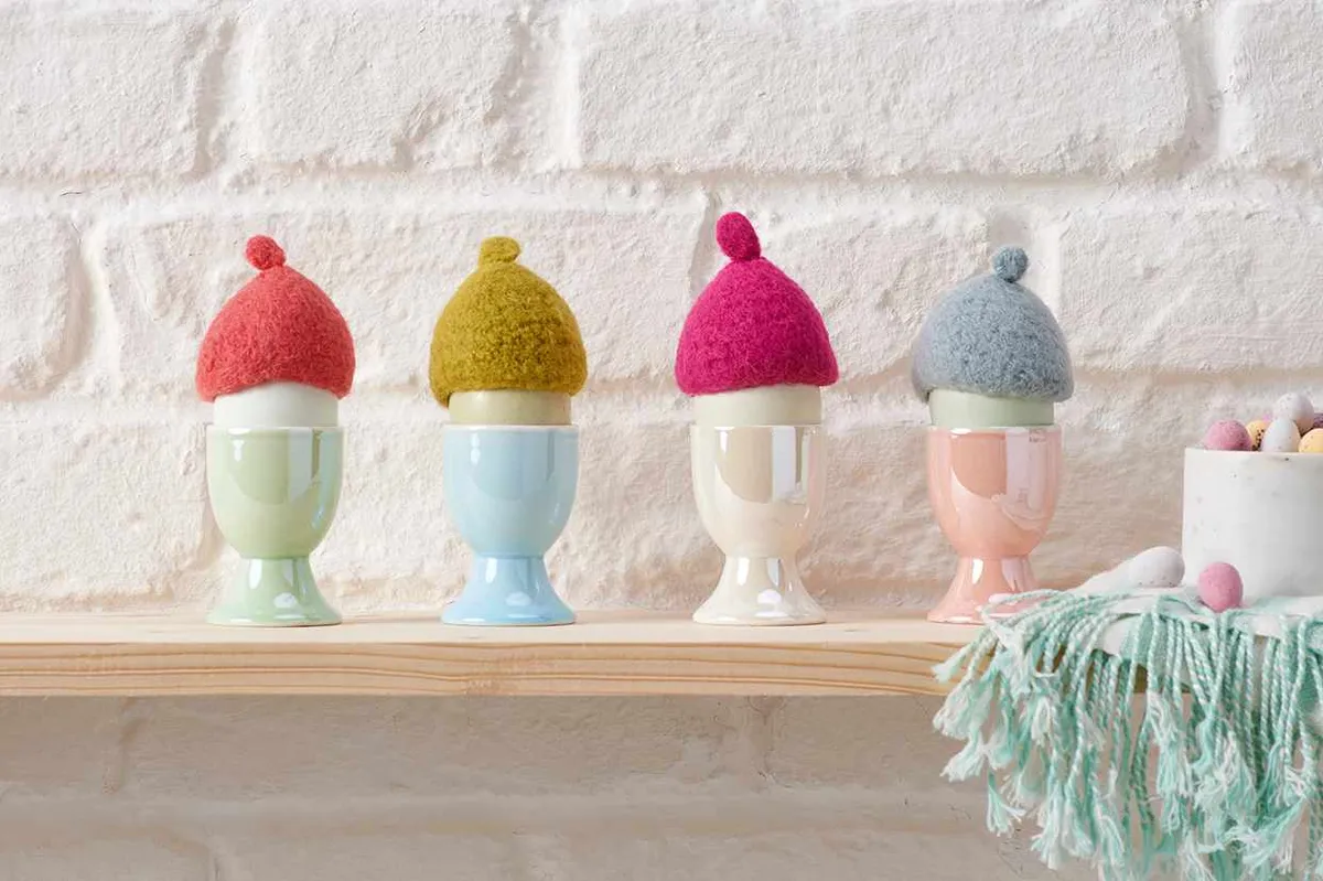 Four mini egg cosies made in different colours of mohair yarn sit on top of 4 boiled eggs in egg cups like they are little bobble hats