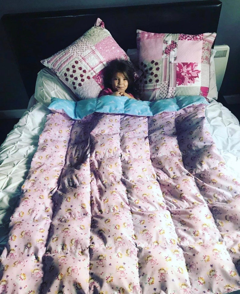 the weighted blanket is spread across a child in bed with a pink patterned fabric on one side and a plain blue fabric on the other side