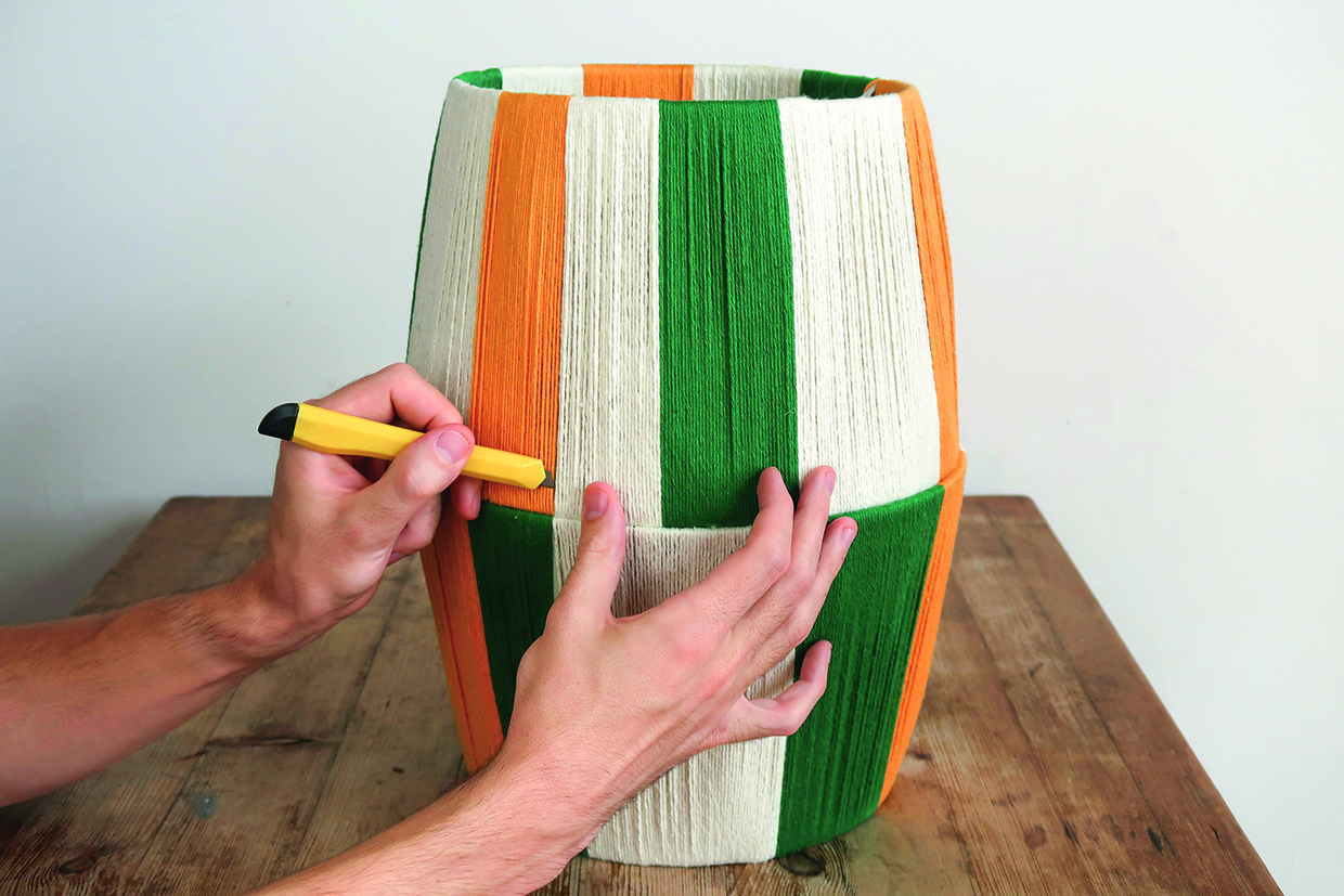 the two yarn lampshades sit stacked on top of each other, with the wider ends facing each other. Hands use a craft knife to cut a slit in the yarn lampshade