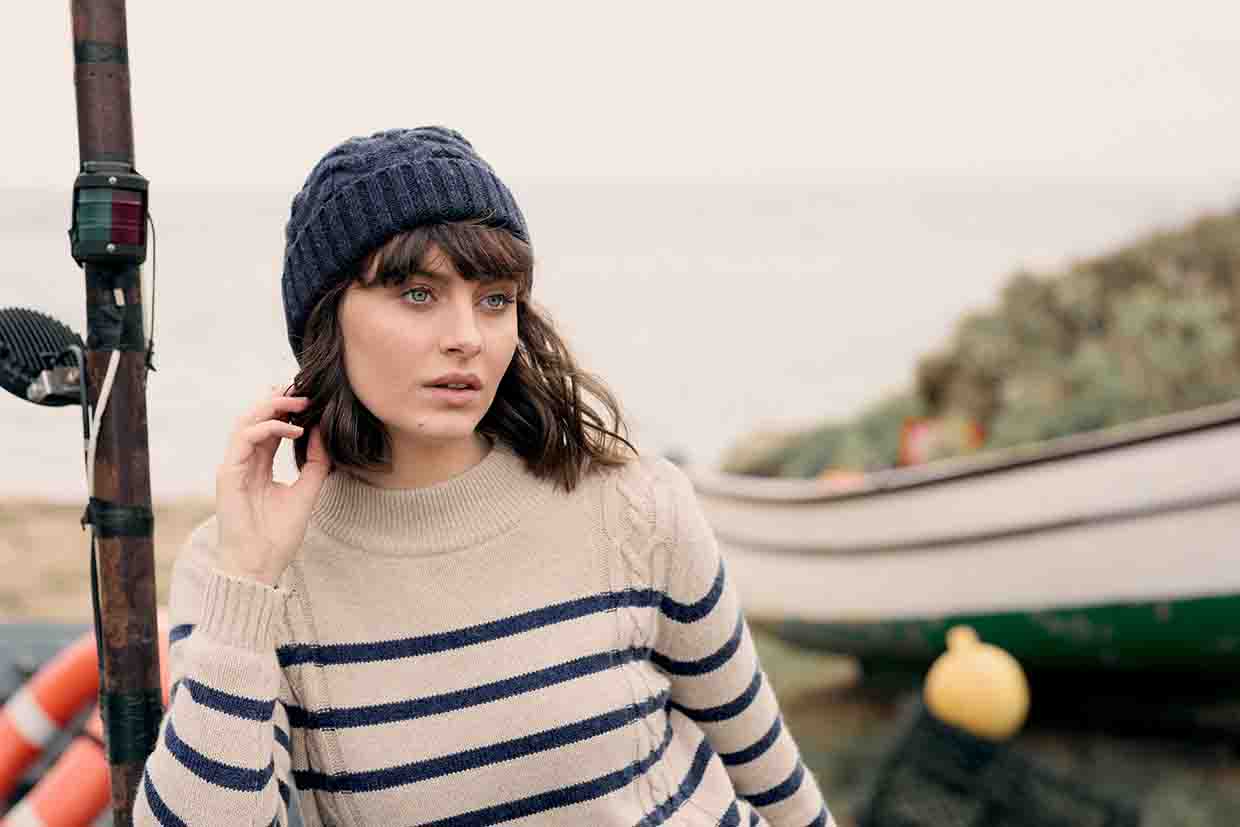 a woman stands inform of a boat on a beach, wearing swollen hat and stripy bable sweater, staring thoughtfully into the distance