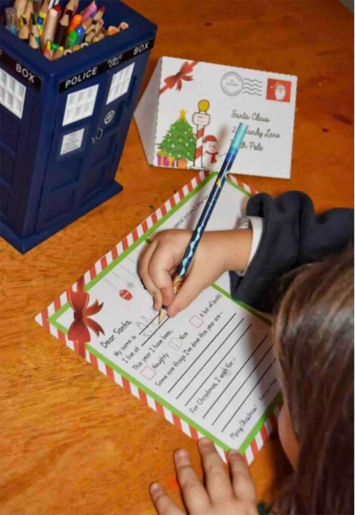 A childwrtites their Christmas letter to sanatorium on a wooden table, with a pot of coloured pencils in a tardis pencil case alongside