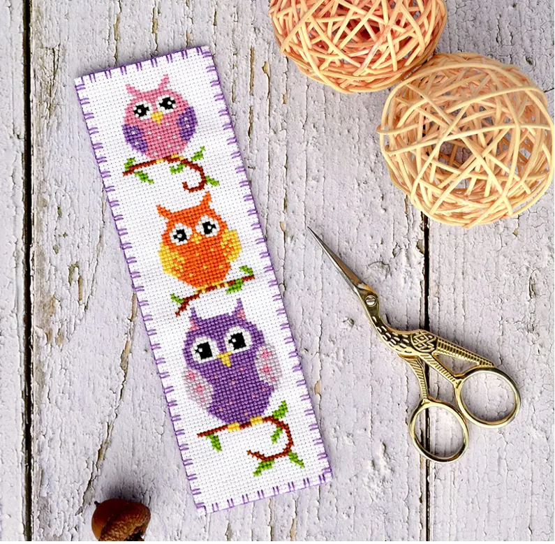 3 owls sit one above each other in a thin line, each arched on a branch. They are cross stitched in purple and orange colours