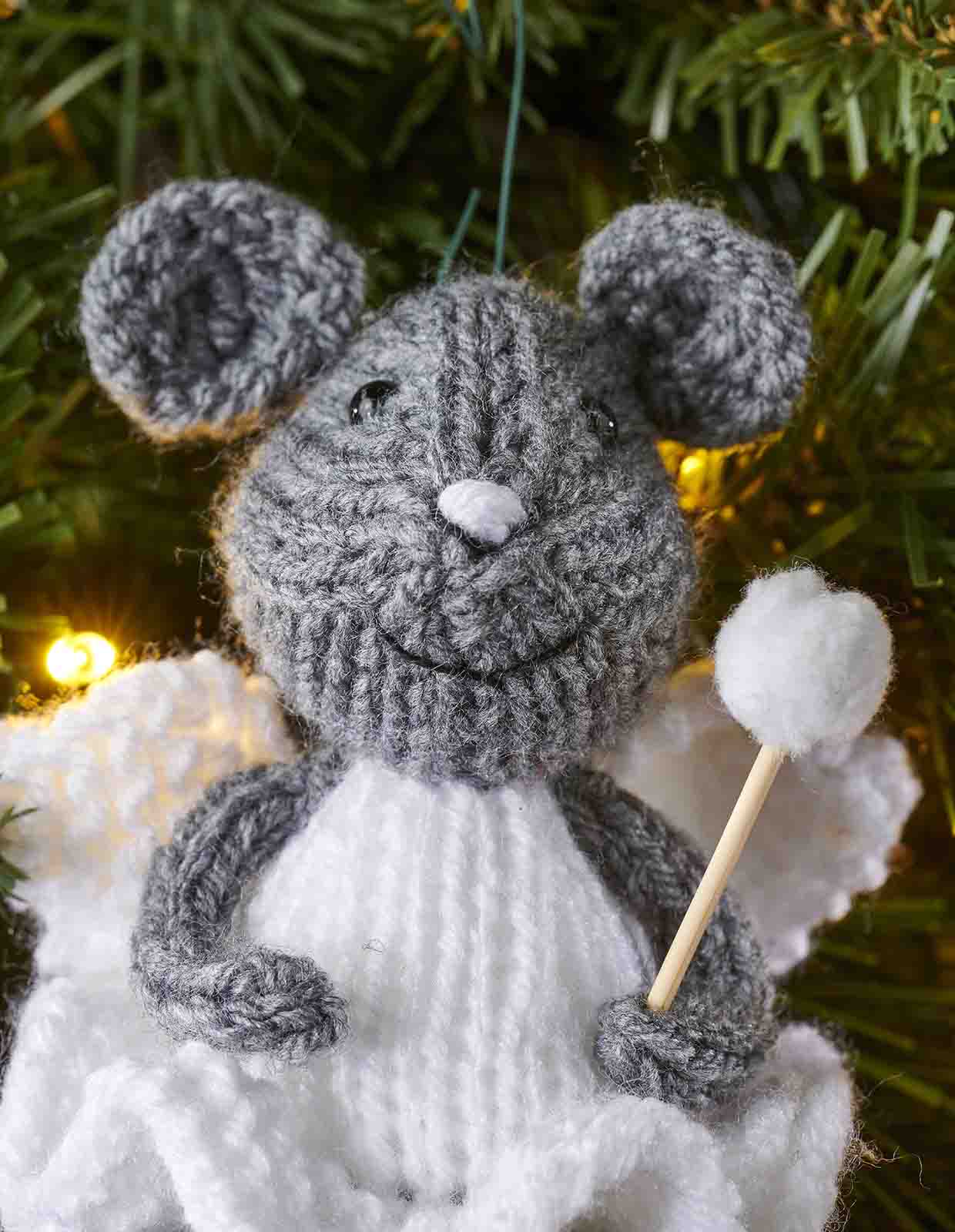 A grey knitted mouse face with two round ears, black eyes, white embroidered nose and smiling embroidered mouth