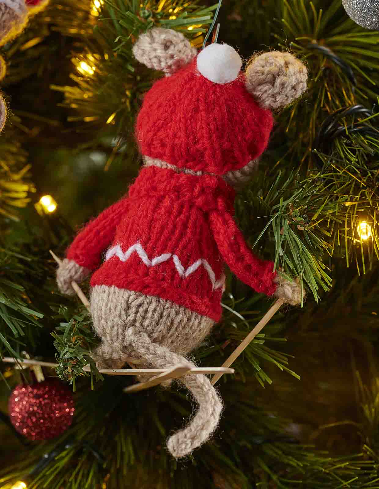 The knitted mouse is seem from behind with his wooden coffee storer skis crossed over under his long icord tail