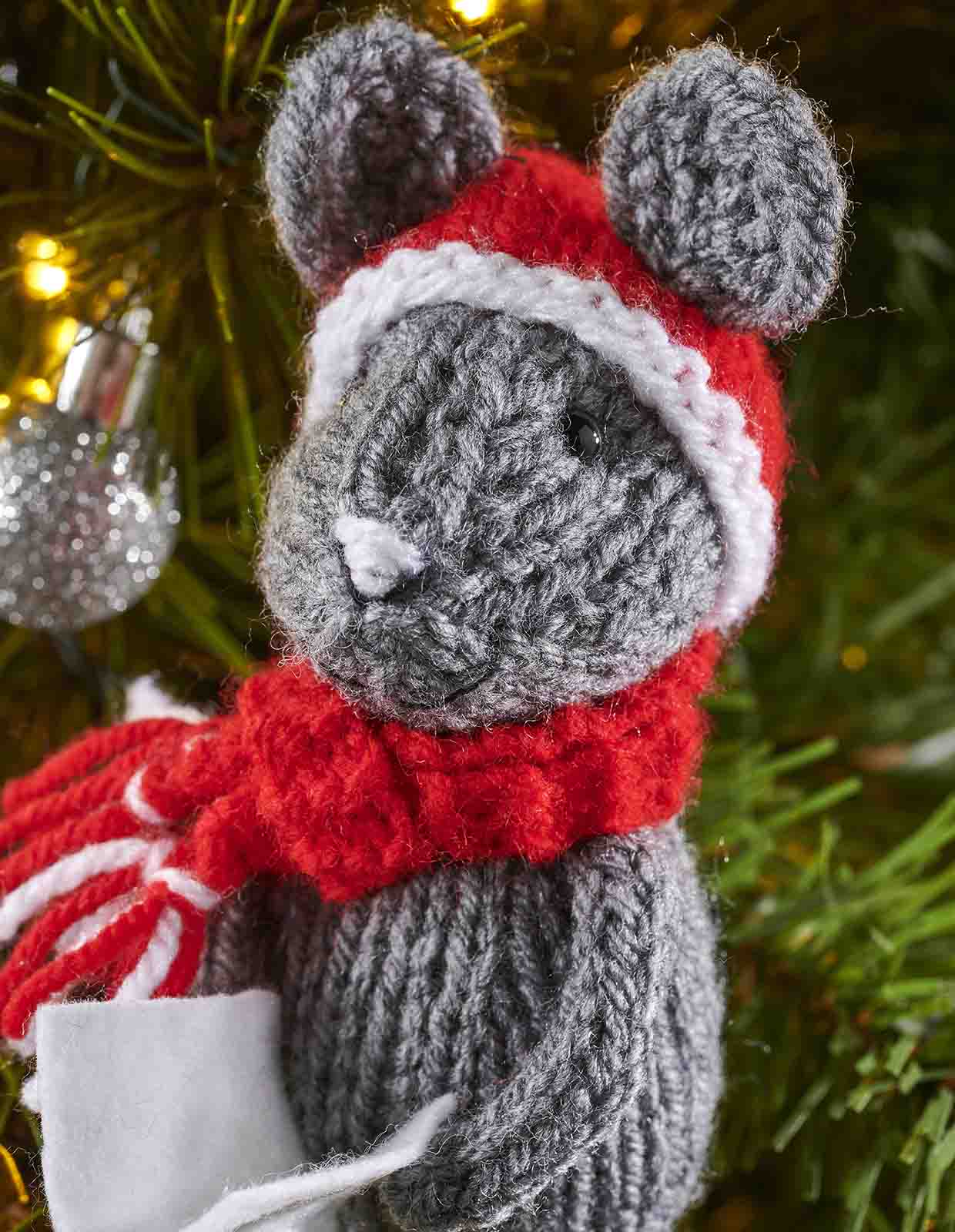 the sweet embroider face of the knitted carol singer mouse is seen close up, the mouse wear a red knitted hat and scarf and looks as thought it is smiling
