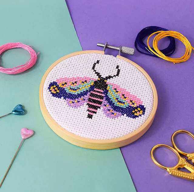 a moth on white fabric cross stitches in green, purple and nblank sits in a hoop on a background of purple and teal with pinks and accessories placed prettily around