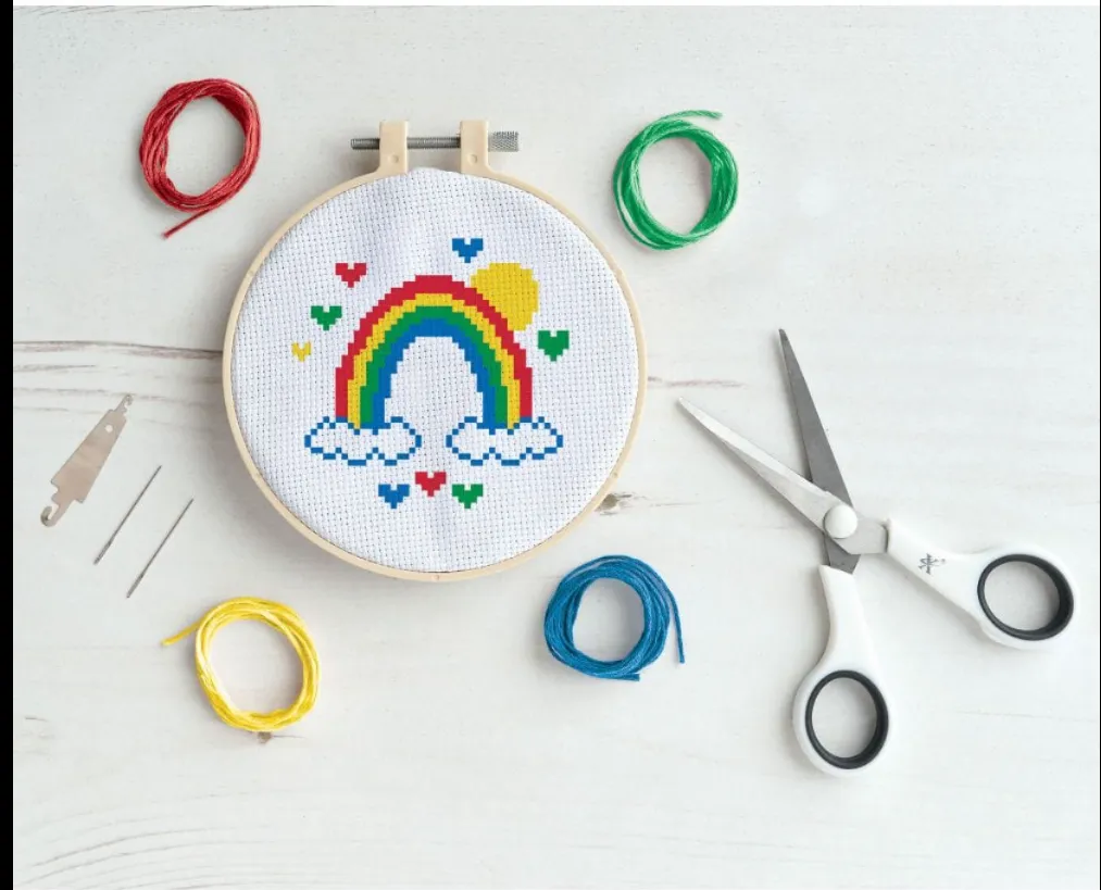 torts od red, green, blue and yellow thread site around a cross stitch design of a rainbow in a hoop that uses those same 4 colours