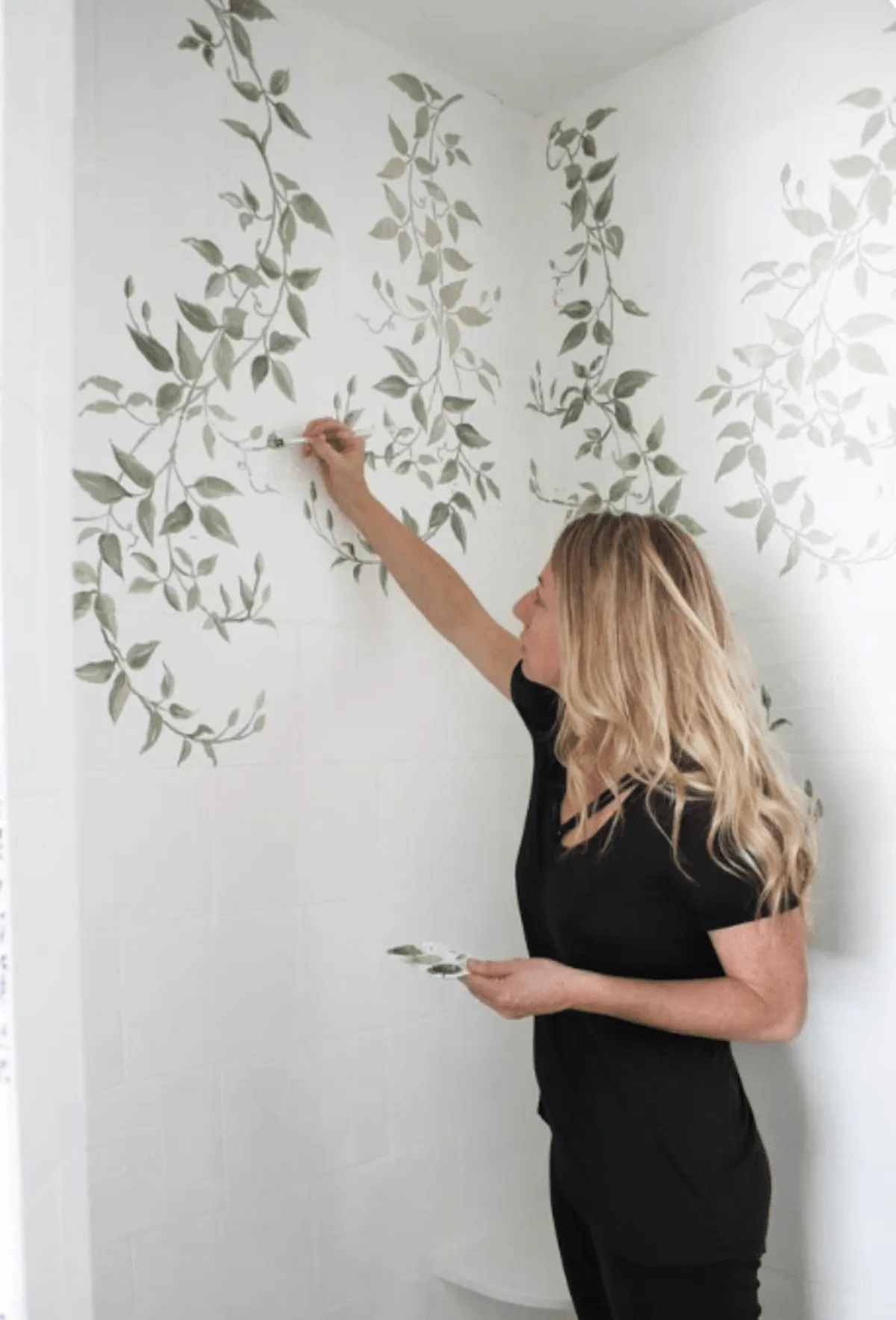 Artist painting a mural featuring foliage and green vines on plain white bathroom wall tiles