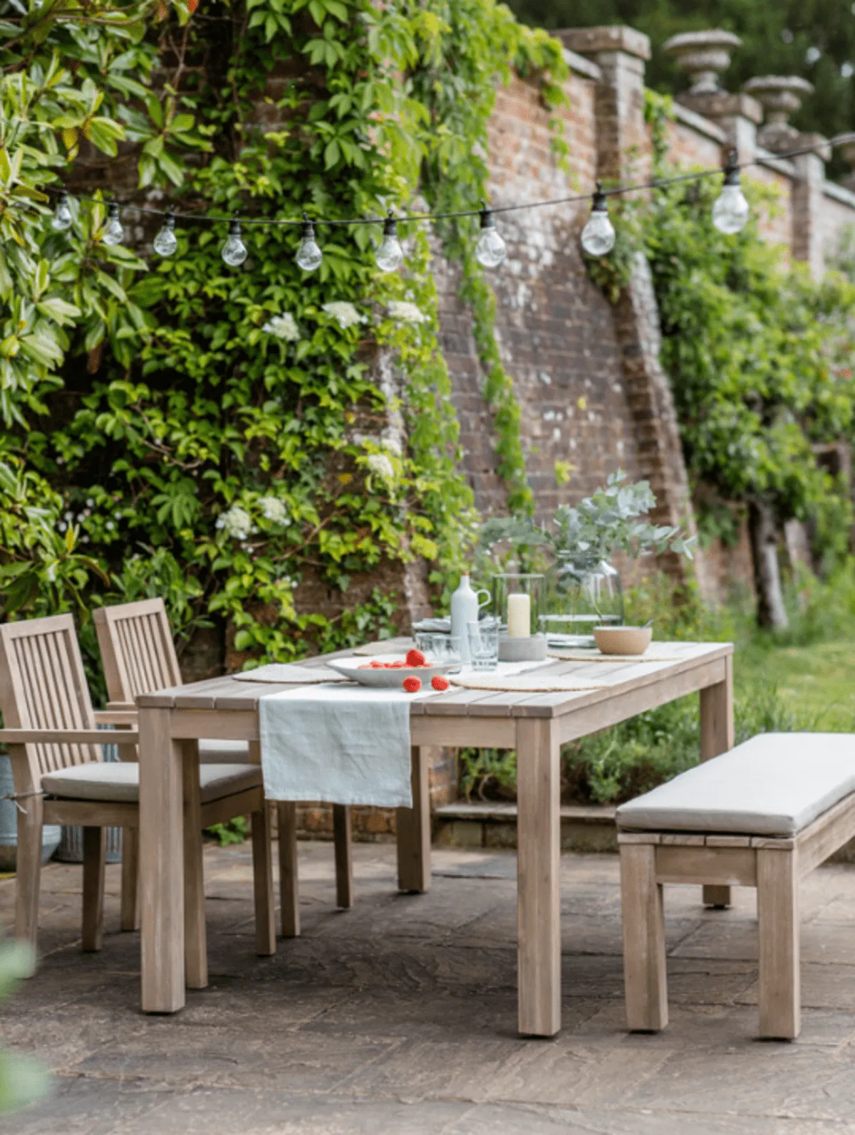 Medium wooden table laid for lunch with bench and two armchairs in a walled garden 