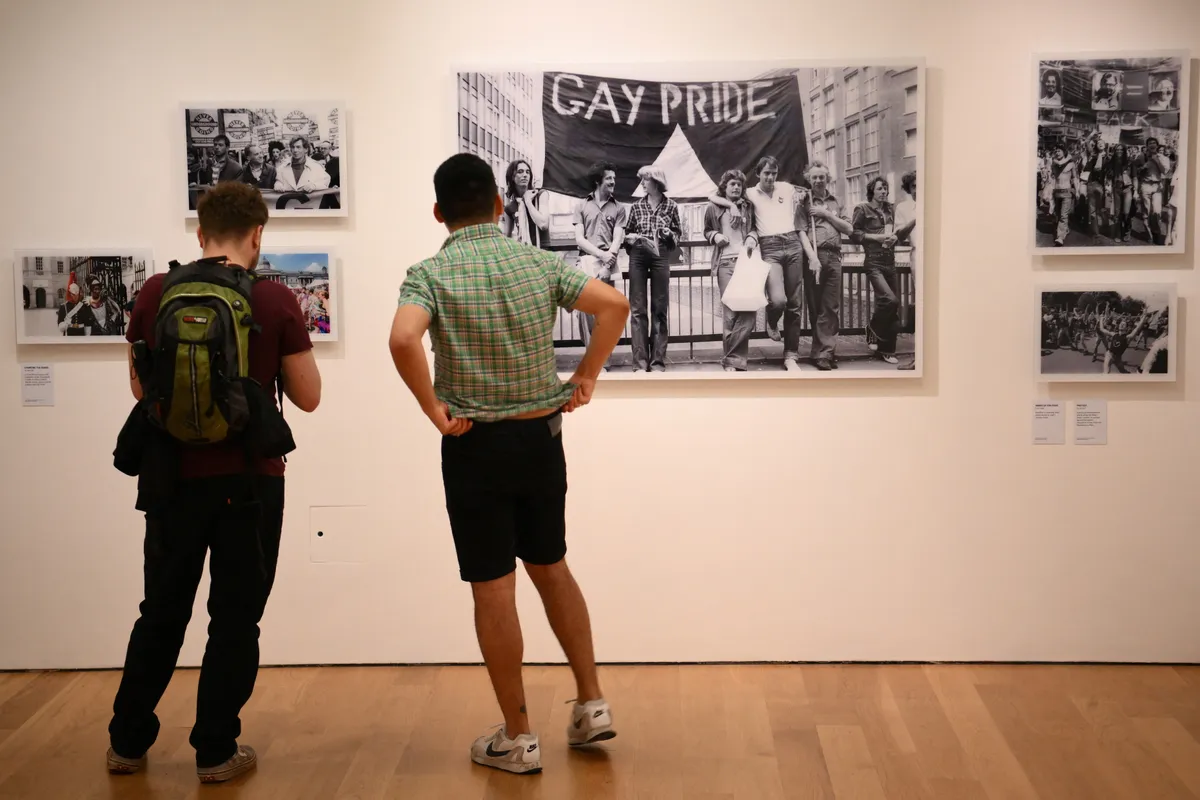 Colour photograph of two white men looking at a black and white photograph of a group of gay rights protesters in the Queer Britain museum