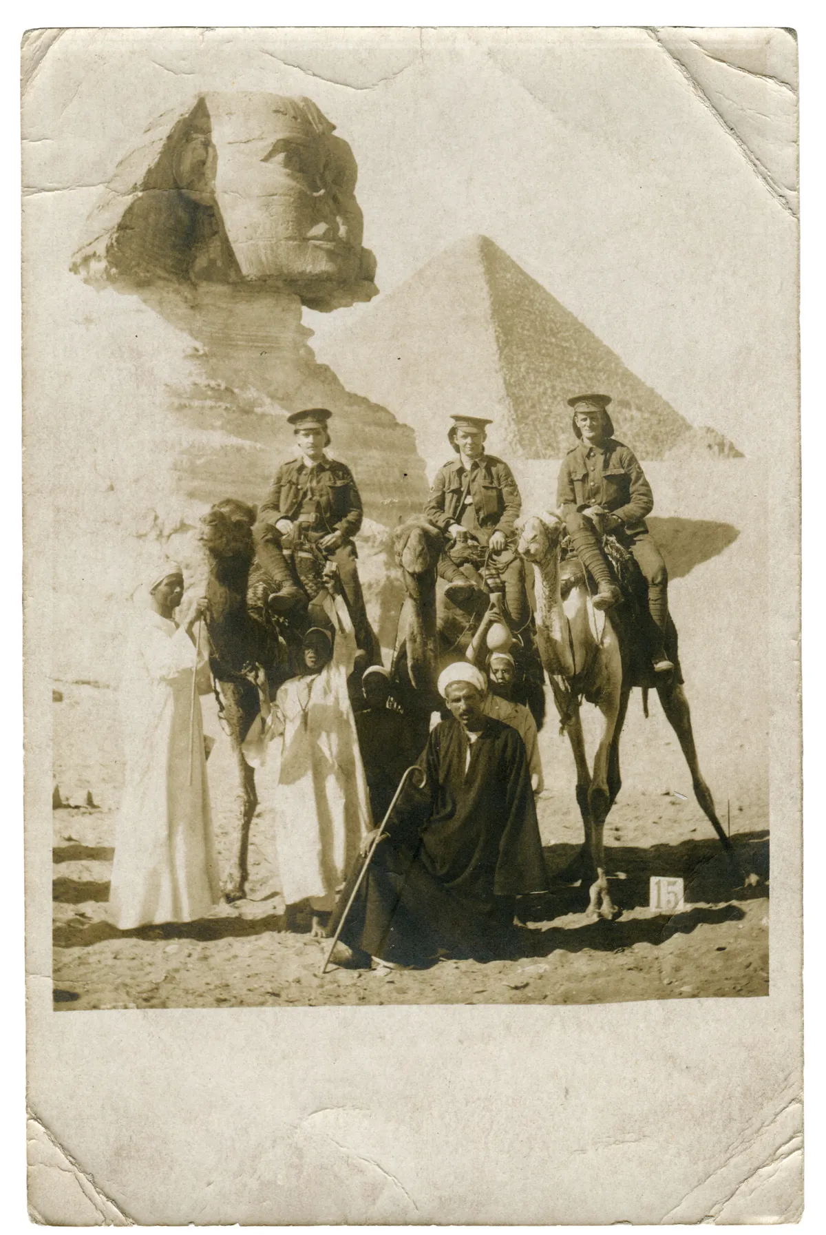 A black and white photograph of three men in British army uniform on camels in front of the Great Sphinx of Giza and a pyramid, with two Egyptian men and two children on foot