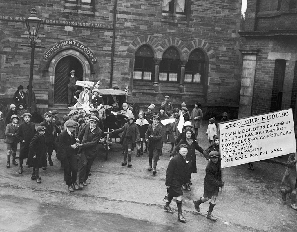 Black and white photograph from the 1920s. A group of men and boys form a procession in the street, pushing a handcart and carrying a banner that says 'St Columb - Hurling'.