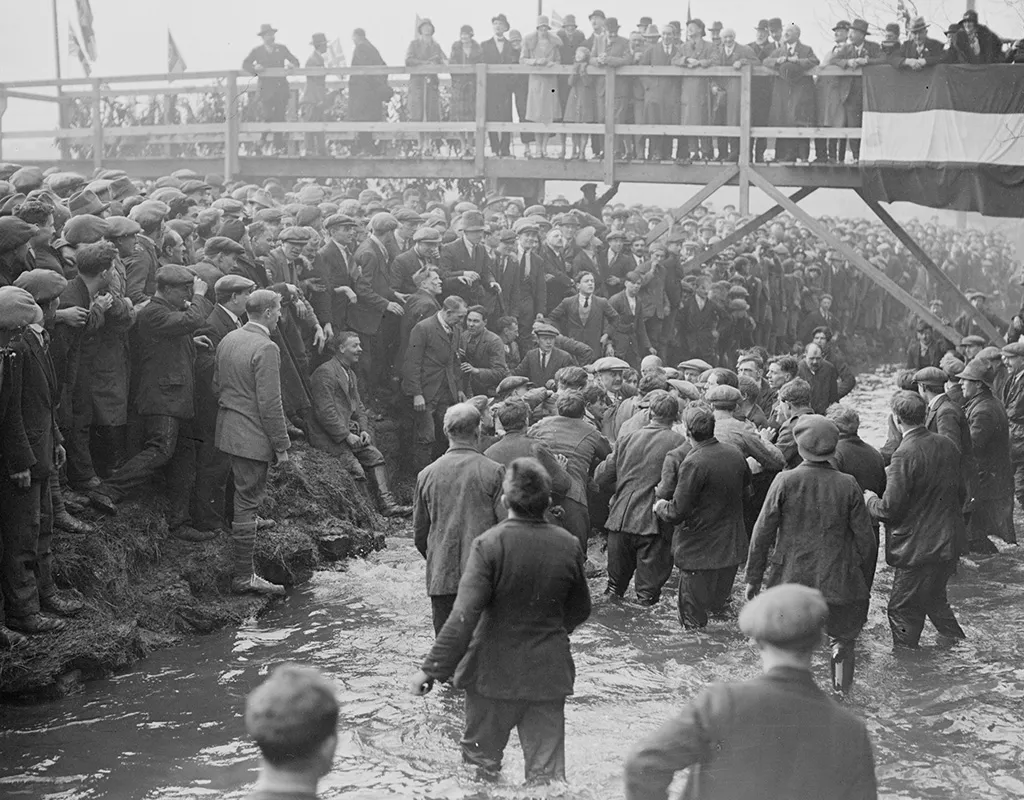 Black and white photograph of a large group of men in 1920s clothing struggling over a ball in a river, while big crowds of men watch from the bank and a bridge overhead.
