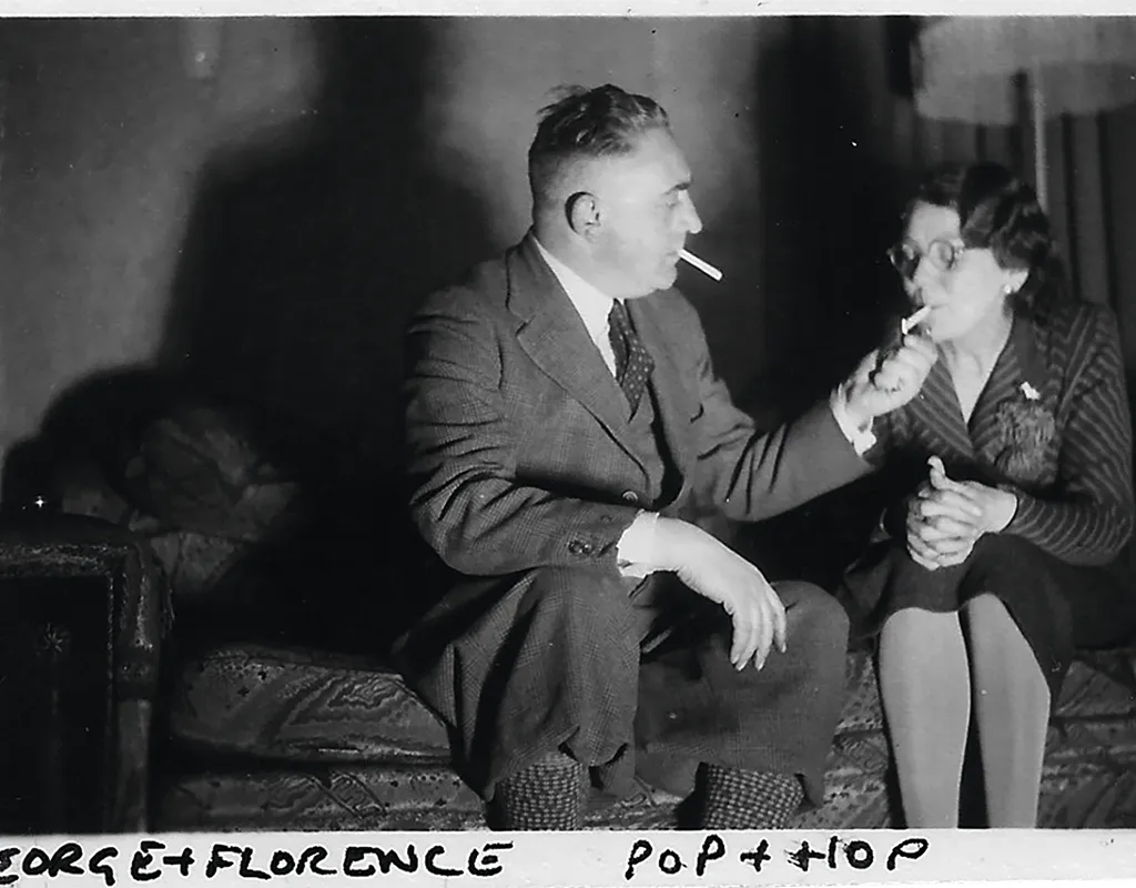 Black and white photograph of a middle-aged white man and woman sitting on a sofa. He's wearing a suit and she's wearing a skirt and dark striped top and glasses. They both have cigarettes in their mouths and he's lighting hers.