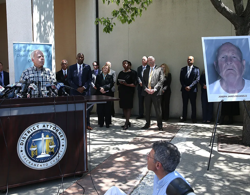 Colour photograph of a man giving a press conference at a podium marked 'District Attorney Sacramento County', with a mugshot of a bald older white man with the caption 'Joseph James DeAngelo' on display next to him