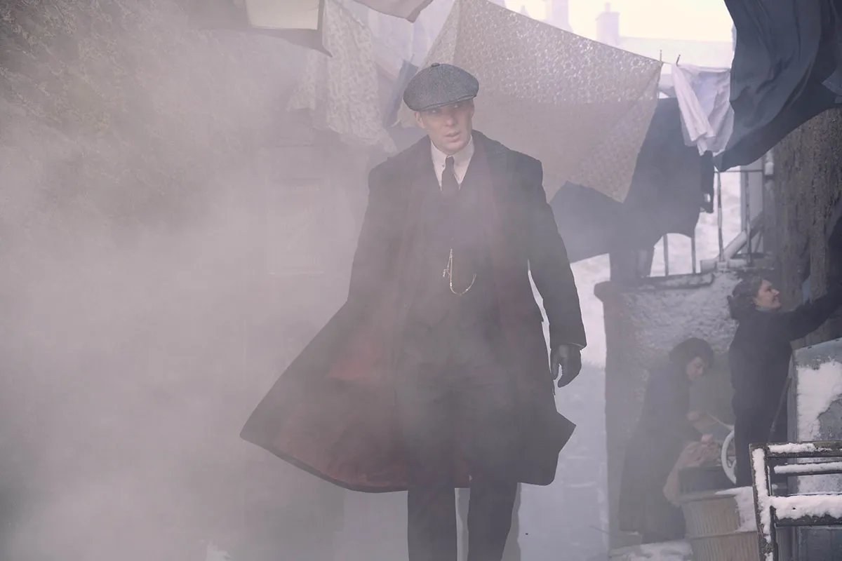A still from the BBC drama Peaky Blinders. Tommy Shelby (Cillian Murphy), a white man wearing a black flat cap and waistcoat and a long black coat, stands in a slum alley with lines of washing strung between the houses.
