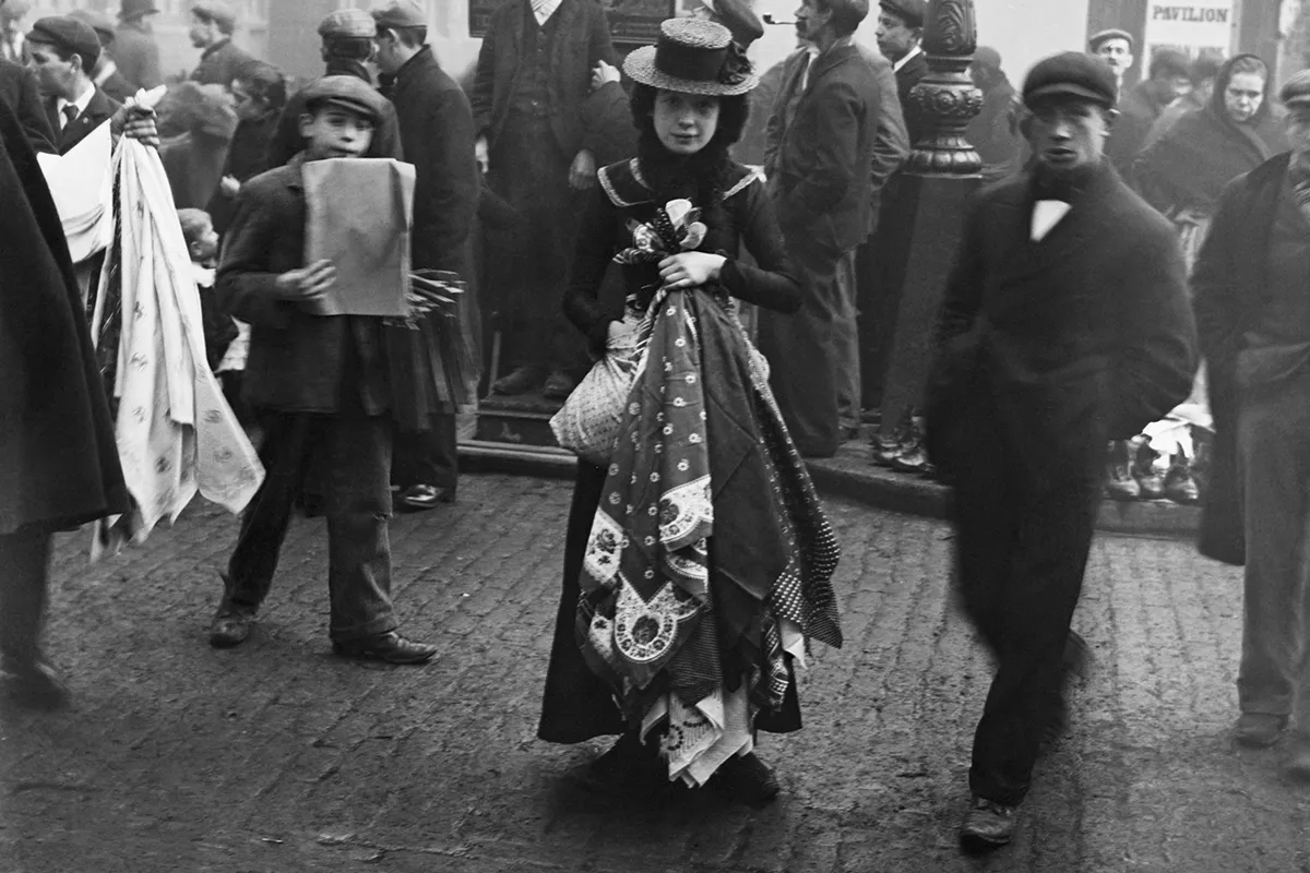 Black and white photograph of a woman in Edwardian dress standing on a street holding a set of handkerchiefs