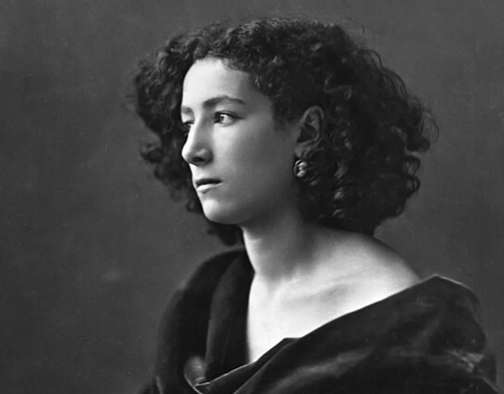 A black and white photograph of Sarah Bernhardt, a 19th century actress. She's a white woman with black curly hair and is wearing earrings and a black gown that exposes her shoulders. She's looking sideways away from the camera with an intense expression.