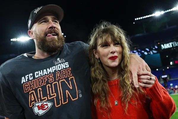 Colour photograph of Travis Kelce, a white man with a brown beard wearing a dark blue American football uniform and cap, and Taylor Swift, a white woman with long blonde hair wearing a red jumper and red lipstick, posing in a football field. He has his arm around her and they're both smiling.