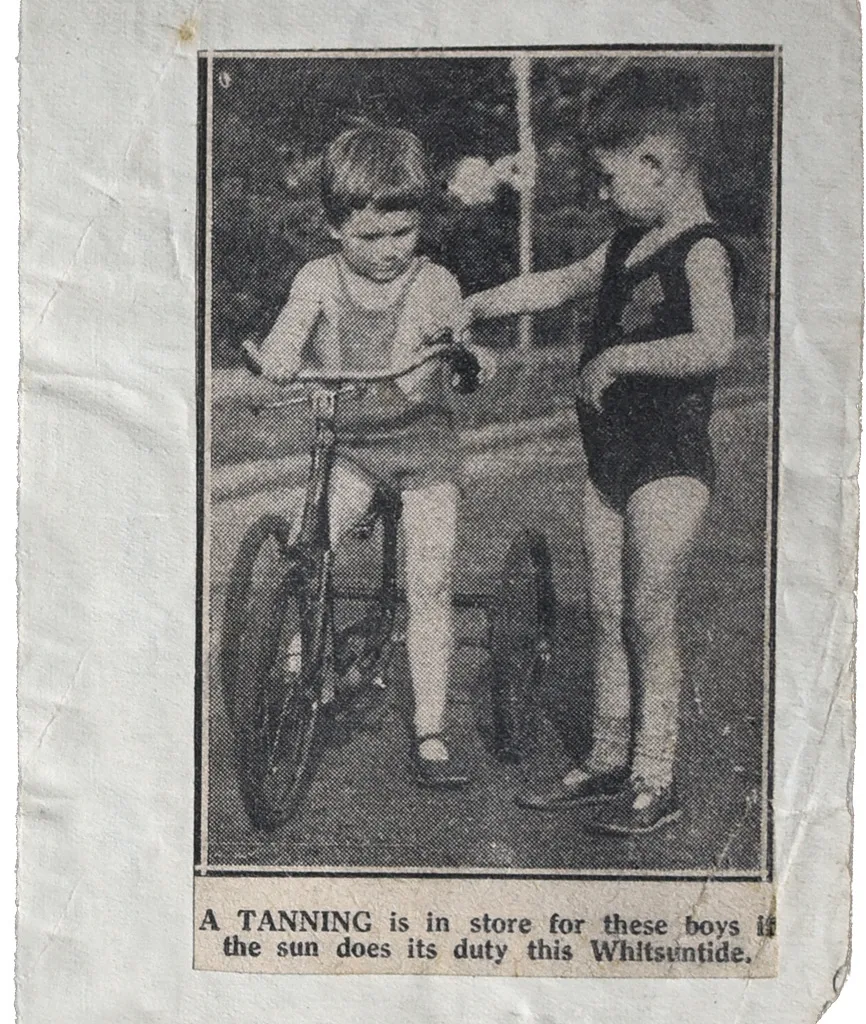A black and white newspaper photograph of two white little boys in swimming costumes. The one on the left is riding on a tricycle.