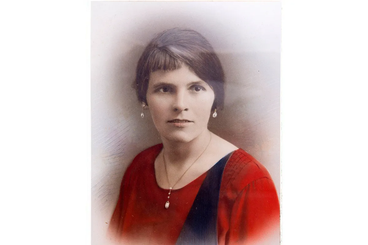 Colourised photograph from the 1920s of a white woman with brown hair wearing a red dress and pearl earrings and a pendant