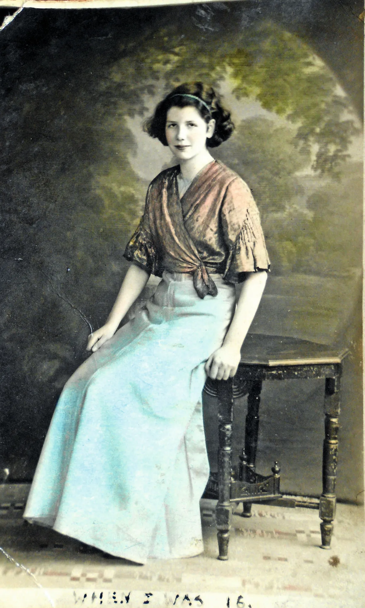Colour-tinted photograph of a young woman with brown hair wearing an orange shawl and long blue skirt.