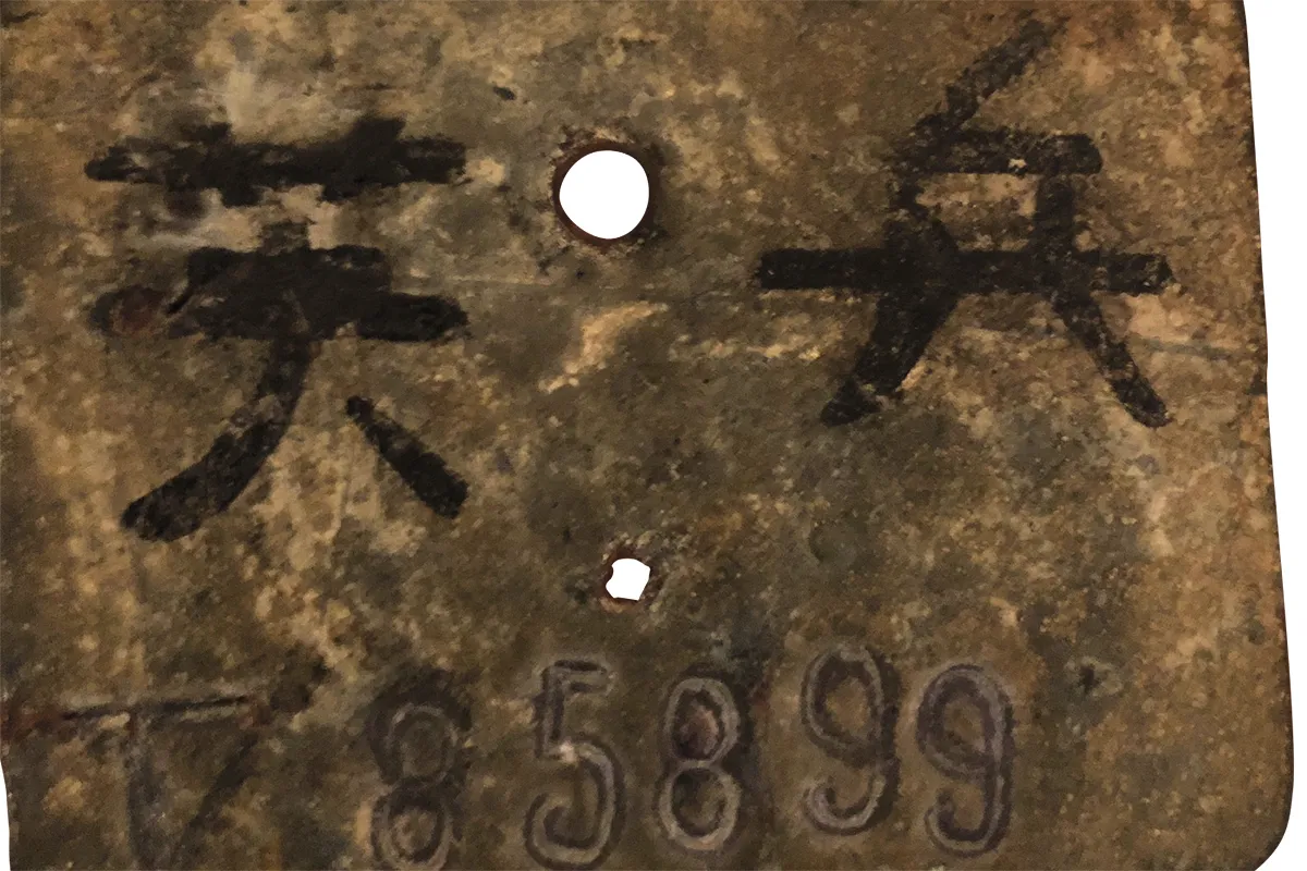 A rusty metal dog tag, with two Japanese characters carved on it and beneath them 785899 in Roman numberals