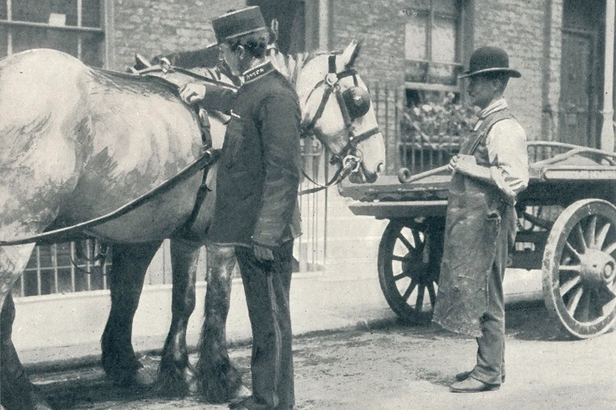 Black and white photograph of a man in a dark uniform patting a white horse attached to a cart, while another man in a bowler hat looks on