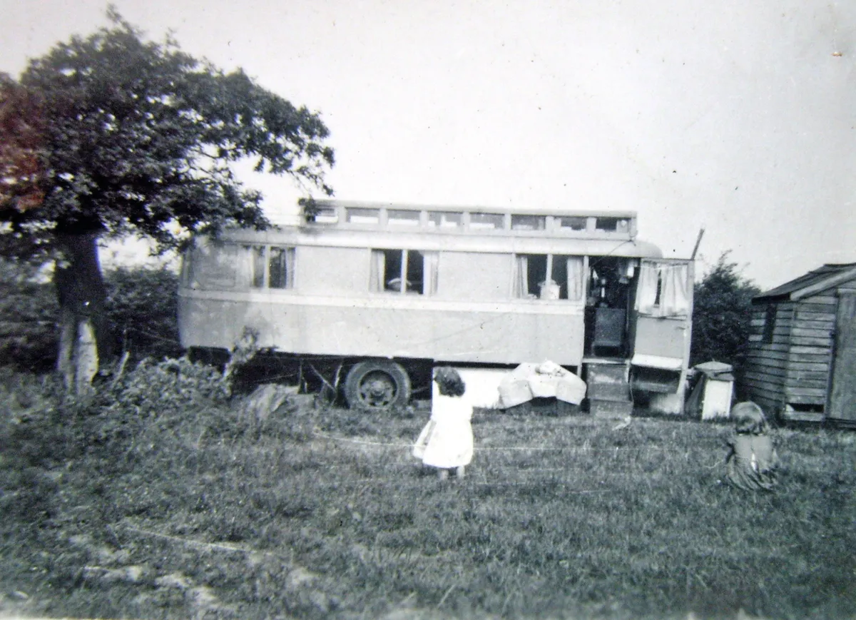 Black and white photograph of an old bus in a field with a little girl standing outside it with her back turned to the camera