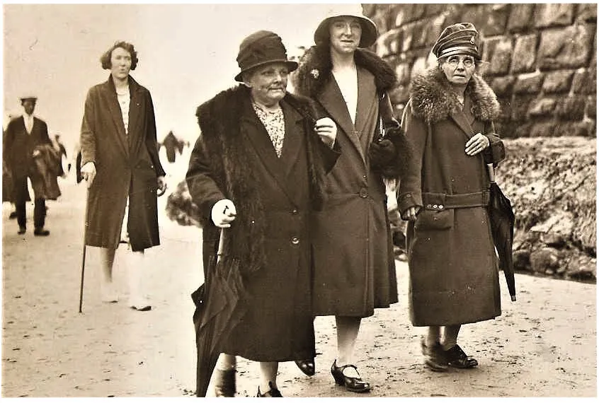 Black and white photograph showing a young woman in Edwardian dress arm-in-arm with two older women and walking along a beach