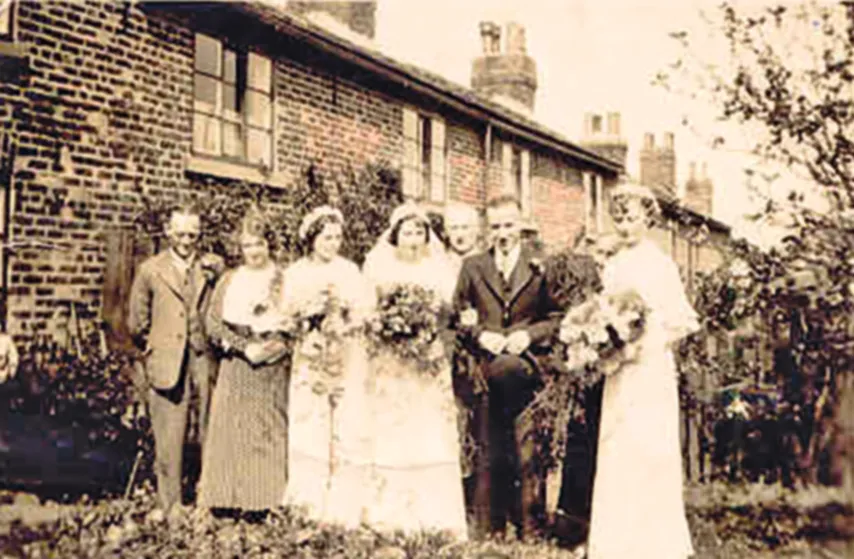 Black and white photograph of a bride posing outside a cottage with her bridesmaids and others.