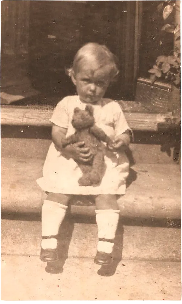 Black and white photograph of a toddler in a white dress holding a teddy bear