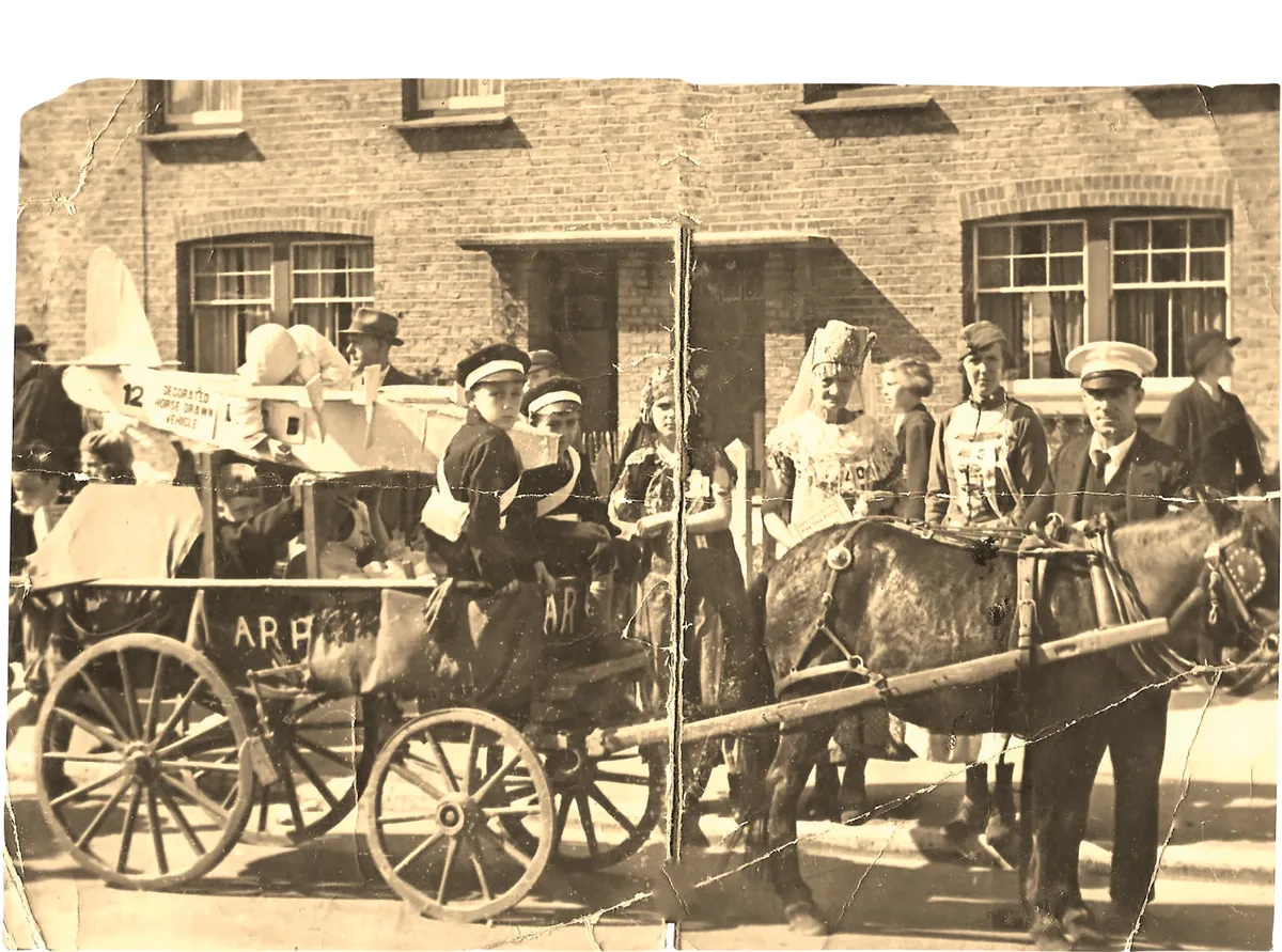 Black and white photograph of a group of children riding a pretend milk cart and horse while some adults stand around