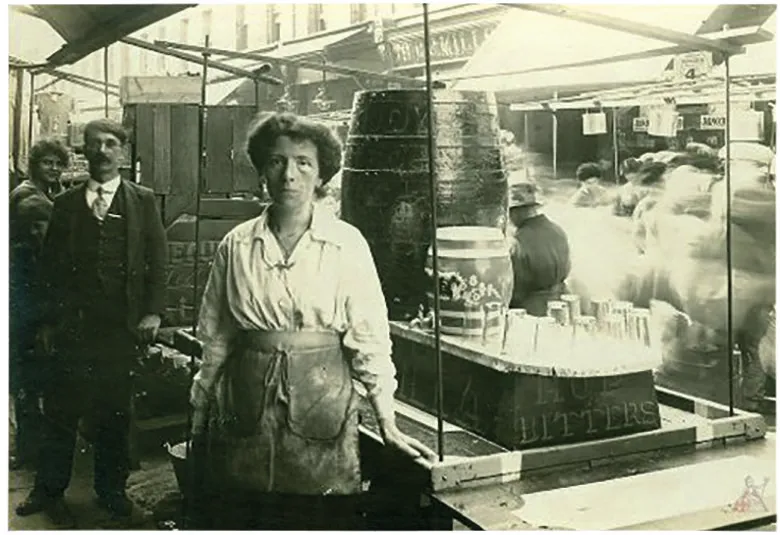 Black and white photograph of a woman standing at an East End market stall with a large barrel
