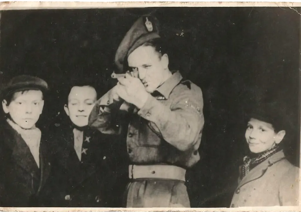 Black and white photograph of a man in Second World War army uniform shooting a rifle while three little boys look on