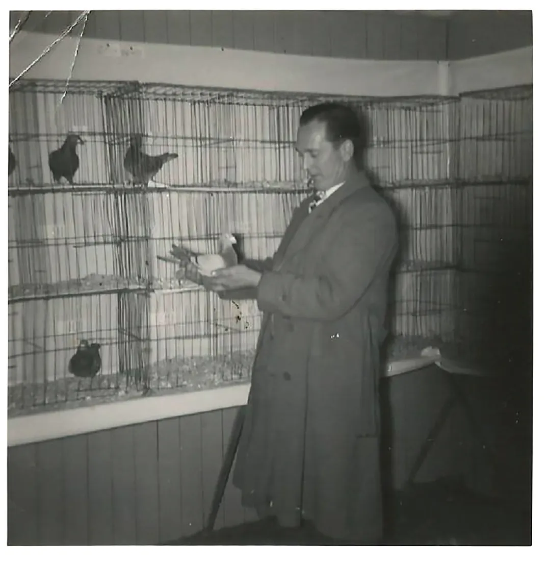 Black and white photograph of a man in a trench coat standing in front of some cages of pigeons