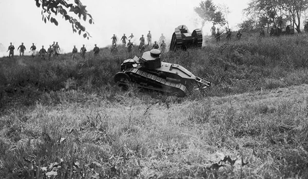 Black and white photograph of a tank crawling over uneven grassy ground with First World War soldiers running behind it