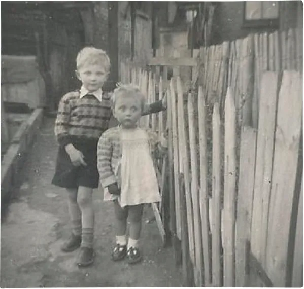Black and white photograph of a little boy and girl in 1950s clothes standing by a fence
