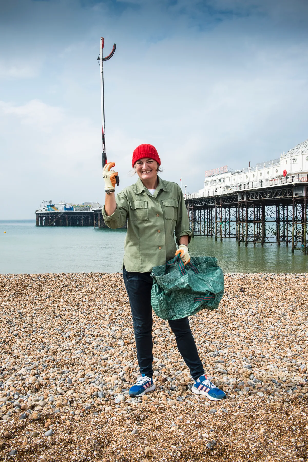 Zoe Lyons in red hat standing in front of Brighton pier holding a litter picker up in the air