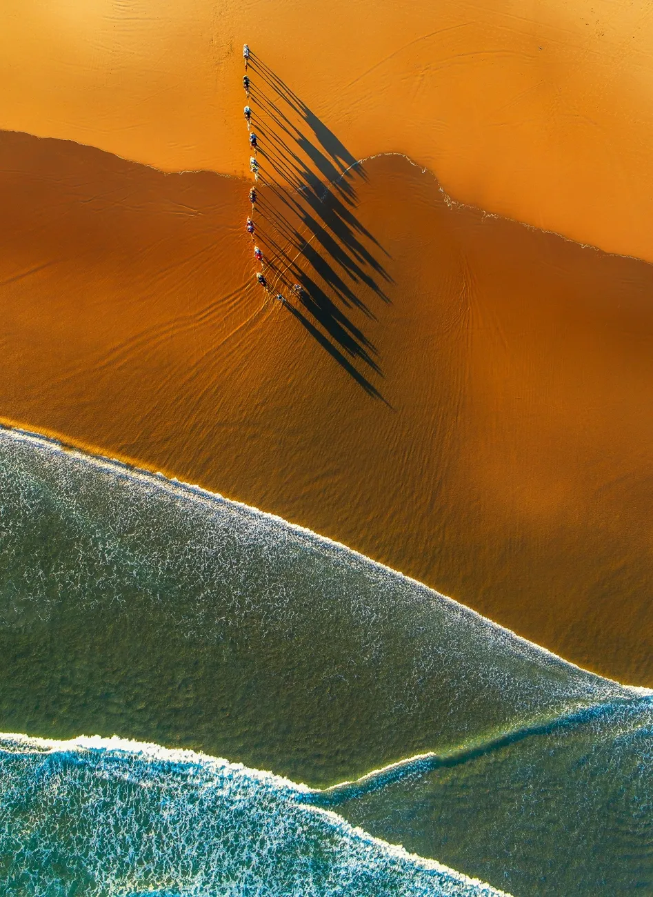 Aerial photo of a desert beach, with water breaking onto deep orange sand and a line of camels, being ridden by people, casts shadows.