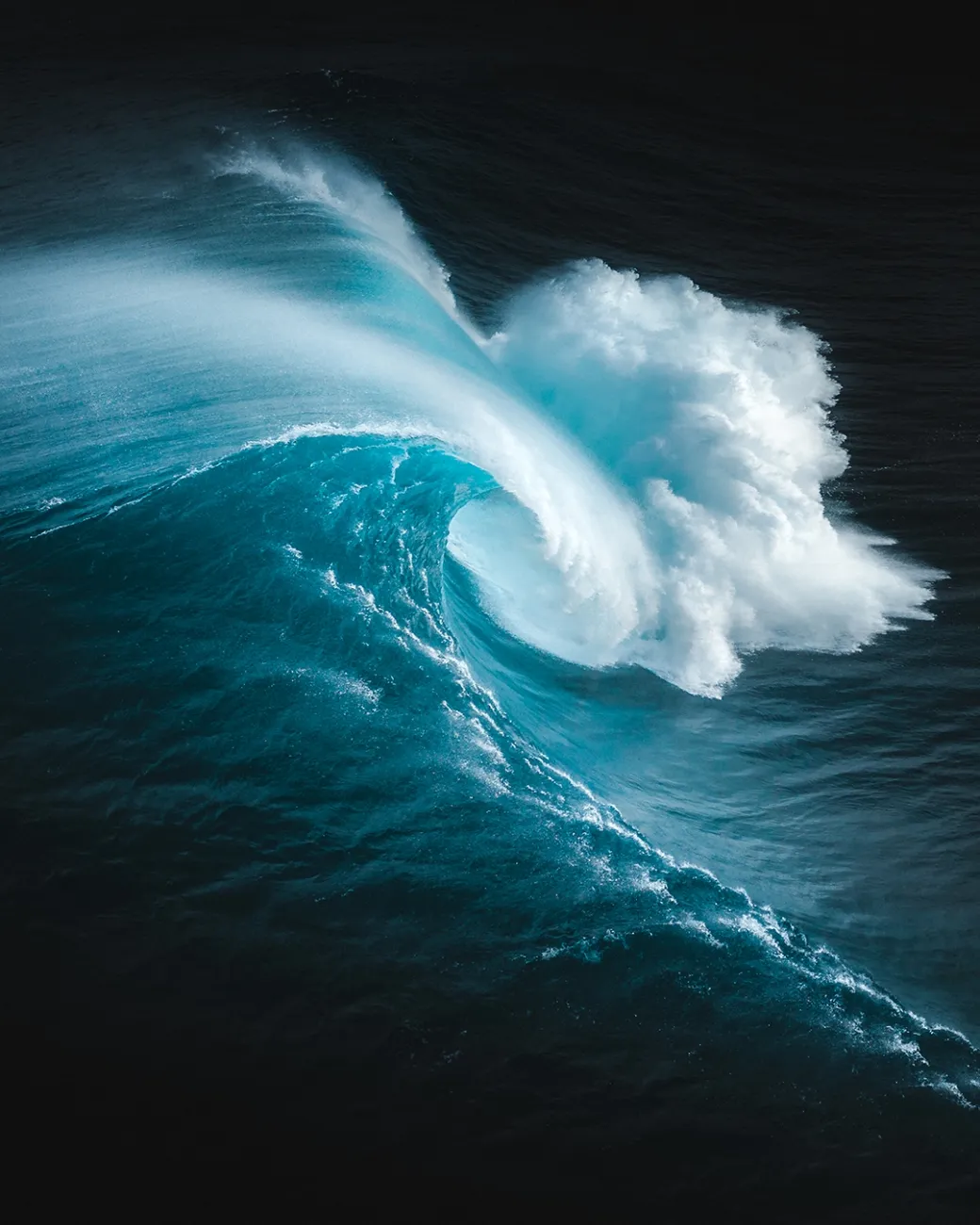 A huge dark blue wave fills the frame, moving from left to right. The edges of the wave are white and curling downwards, crashing into the surface of the water below.