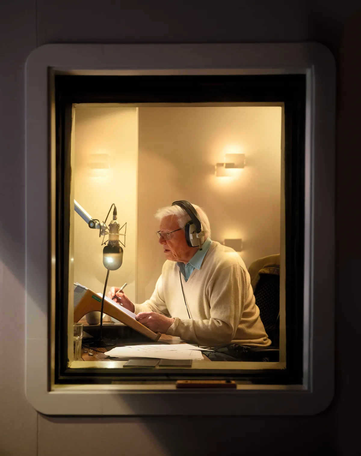 A picture of David Attenborough taken through a window as he is speaking into a microphone with headphones on.