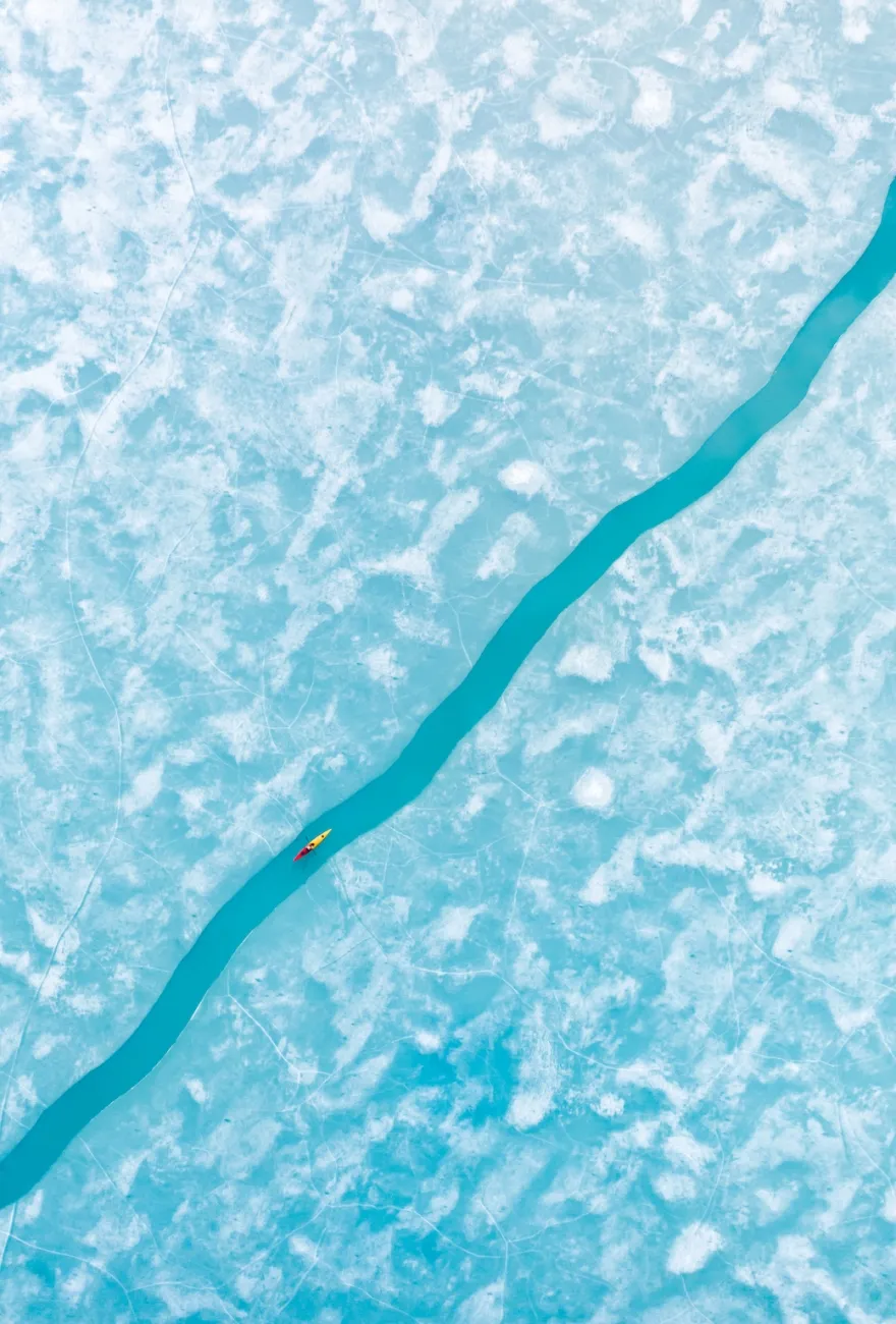 Aerial photo of a blue-white ice sheet, dissected by a thin almost-straight line of blue water running diagonally from bottom left to top right, with yellow and red kayak moving along it.