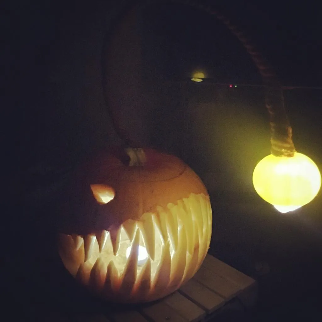 A pumpkin in darkness, lit up from within to show the sharp teeth of an anglerfish carved into the pumpkin, with an additional very bright light hanging in front of the pumpkin.