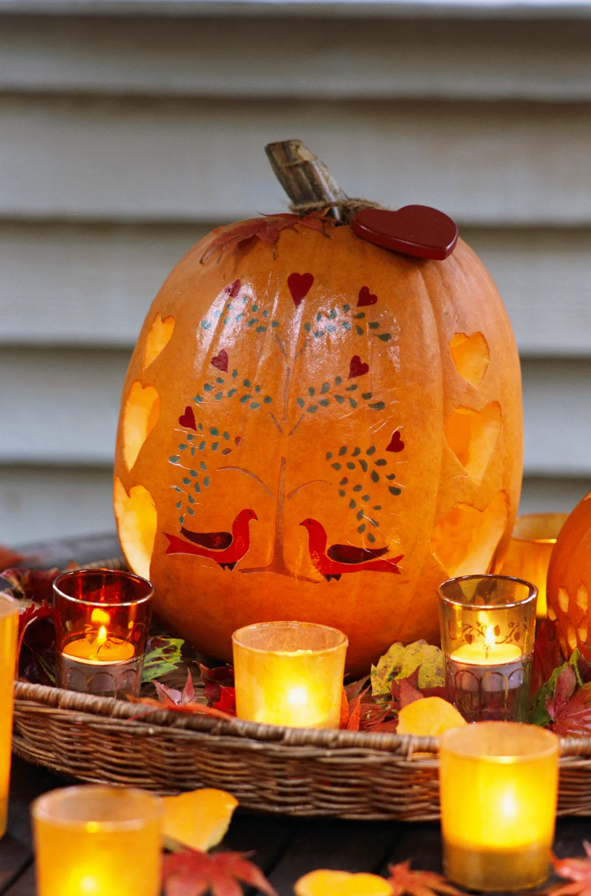 A carved and painted pumpkin featuring painted birds and a tree, and carved hearts, surrounded by votive candles and autumnal leaves.
