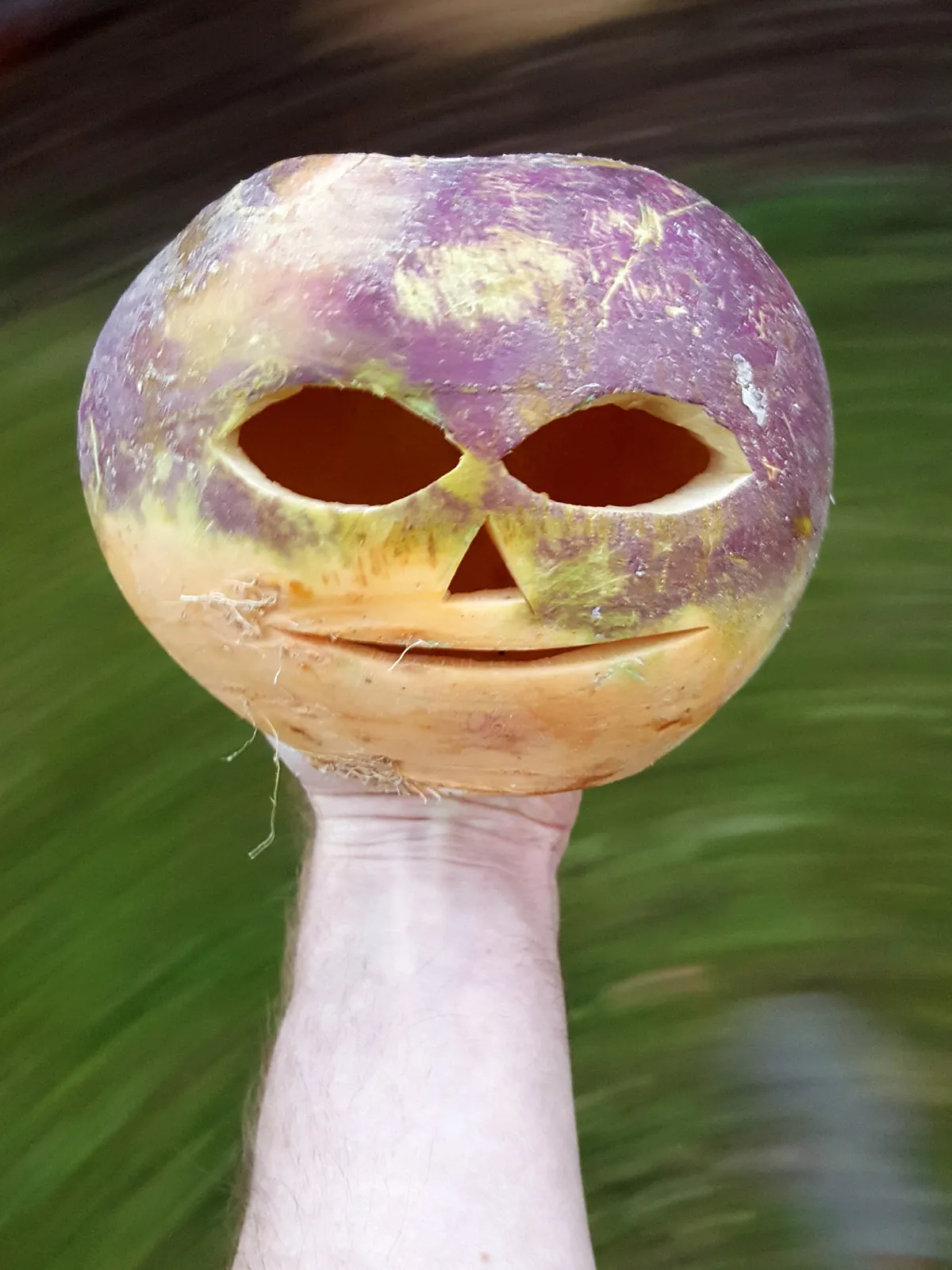 A hand holding a turnip against a blurred background. The turnip has a face carved into it.